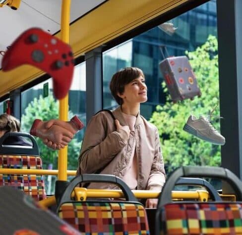 Eine junge Person sitzt entspannt in einem fast leeren Stadtbus und schaut lächelnd aus dem Fenster. Um sie herum schweben verschiedene Gegenstände in der Luft, darunter ein roter Gamecontroller, ein Paar Hände, ein Sneaker und ein gepunkteter Würfel. Die bunten Sitzpolster und der Blick auf grüne Bäume sowie ein modernes Gebäude durch die großen Busfenster sind zu sehen.