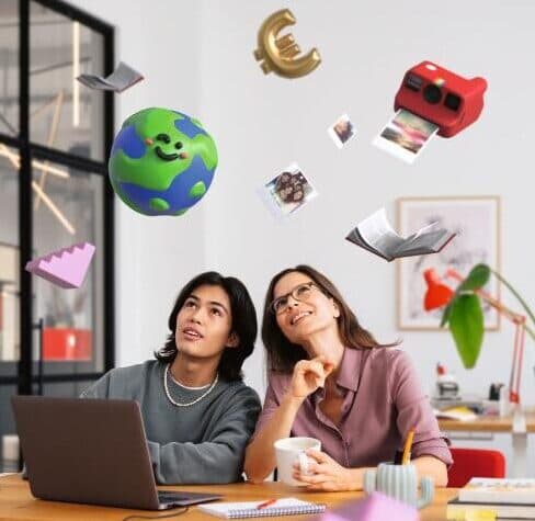 Zwei junge Personen sitzen gemeinsam an einem großen Holztisch in einem modernen, hellen Büro. Vor ihnen liegen ein Laptop, Notizbücher und Stifte. Beide blicken fasziniert nach oben, wo animierte Gegenstände – darunter eine fröhlich lächelnde Erdkugel, das Euro-Zeichen, ein rotes Fernglas, ein Buch und Fotos – scheinbar schwerelos in der Luft schweben. Im Hintergrund sind eine große Pflanze, Regale und moderne Büromöbel zu sehen.