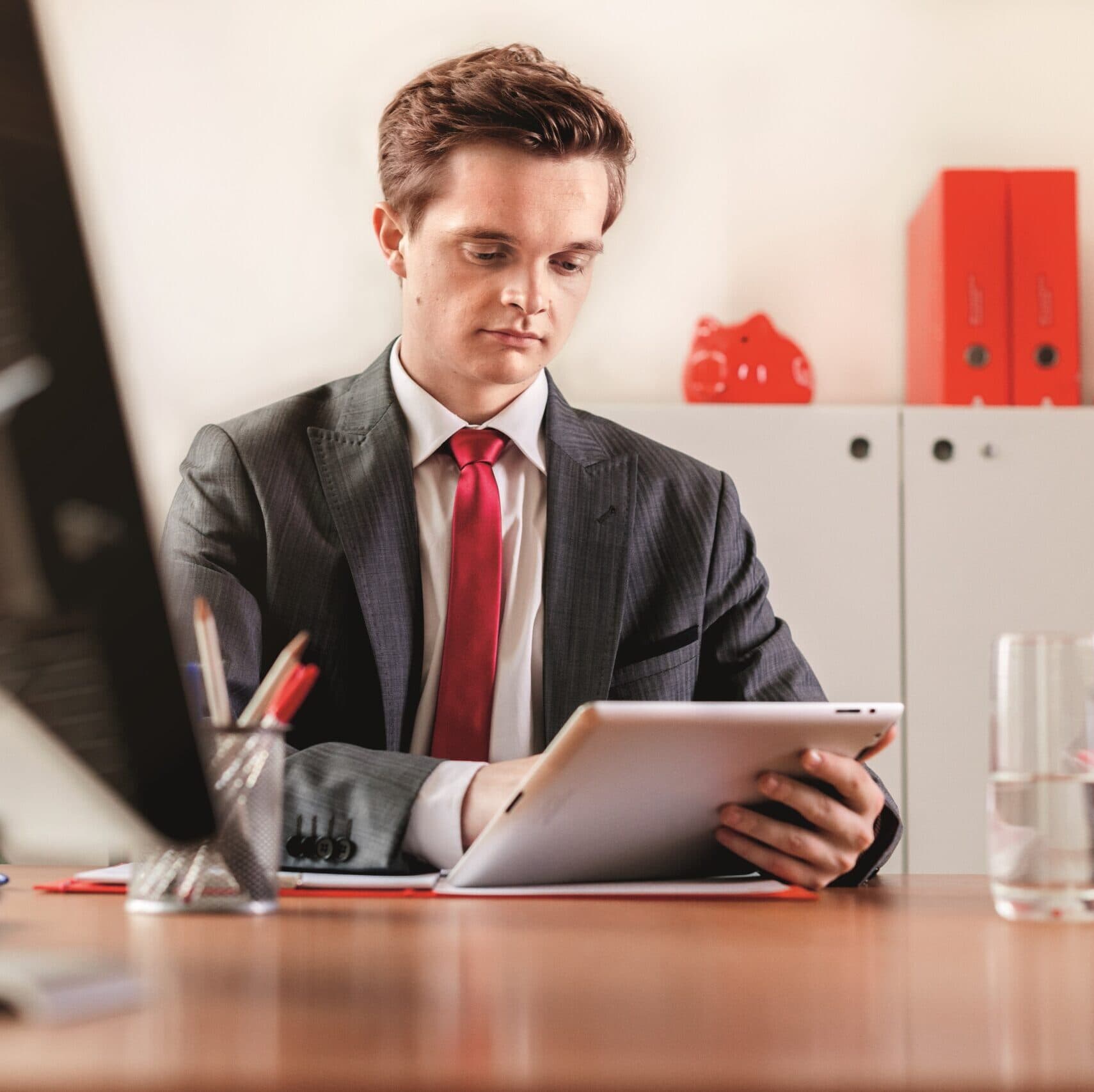 Ein junger Mann in einem dunkelgrauen Nadelstreifenanzug und weißem Hemd mit roter Krawatte sitzt an einem Holzschreibtisch in einem Büro und schaut konzentriert auf ein Tablet in seinen Händen. Auf dem Schreibtisch befinden sich ein Stiftehalter, ein Glas Wasser und Dokumente. Im Hintergrund sind weiße Aktenschränke mit roten Ordnern und einer roten Dekofigur zu sehen.